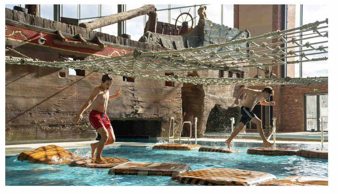 A lively pool area features a pirate-themed structure with floating platforms, where two individuals are engaged in playful activity. Sunlight streams through large windows, highlighting the water's vibrant blue hue and the textured surfaces of the poolside.