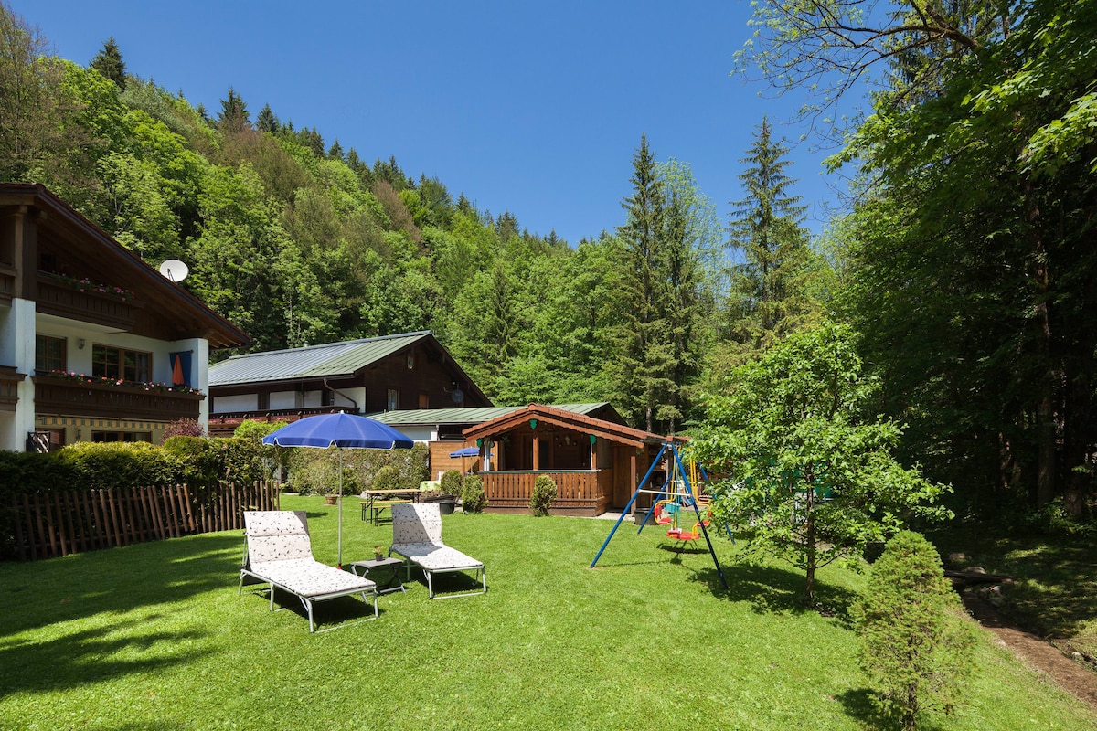The garden area features well-maintained green grass under a clear blue sky. Two lounge chairs are positioned on the lawn, accompanied by a large umbrella. A wooden play structure with swings is visible, alongside a cozy cabin, all surrounded by lush trees.