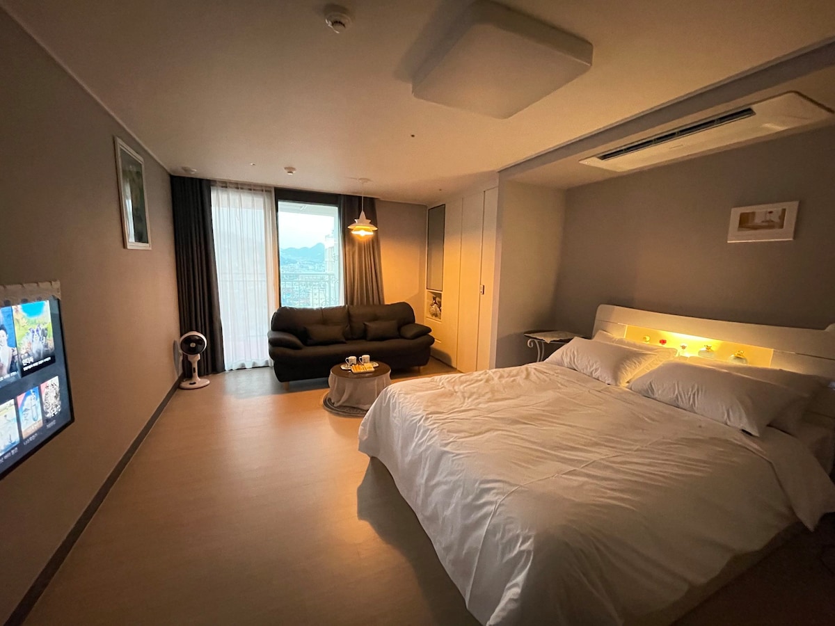 A spacious room is presented with a large bed featuring light bedding. A comfortable dark sofa is positioned near a window, offering views of the outside. A small coffee table is placed in front of the sofa, and a wall-mounted TV is visible.