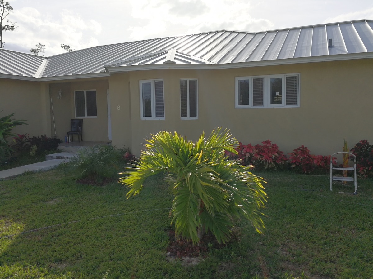 A single-story home is surrounded by lush green grass and vibrant landscaping, featuring a palm tree and colorful flower beds. Light plays on the metal roof, while multiple windows allow natural light to brighten the interior spaces.