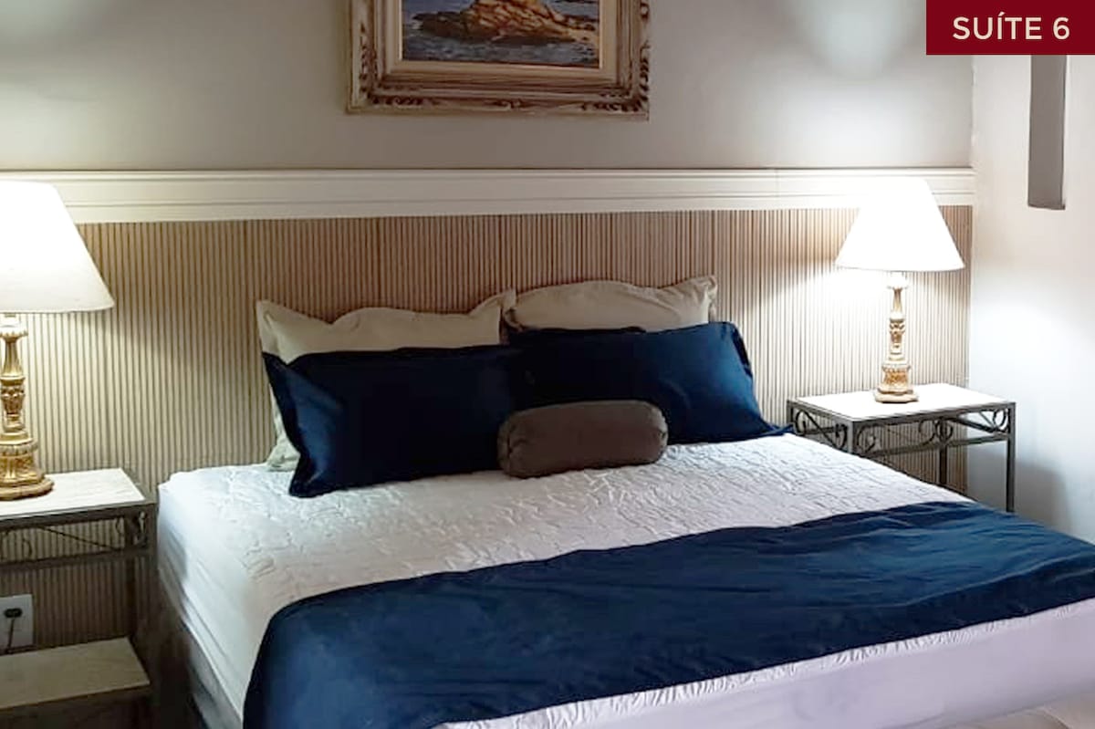 A well-appointed sleeping space features a spacious bed with a white quilt and navy blue accent pillows. Nightstands with lamps are positioned on either side of the bed. A framed artwork adorns the wall above, adding a touch of elegance to the room.
