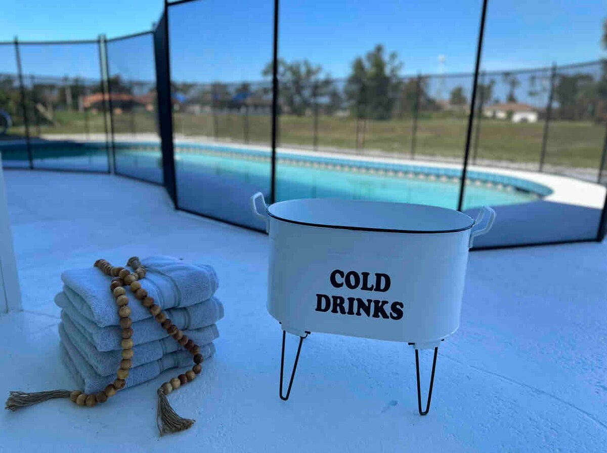 A white container labeled 'COLD DRINKS' is positioned beside neatly stacked gray towels. The background features a blue heated pool enclosed by a safety fence, with a view of green lawns and trees enhancing the outdoor space.