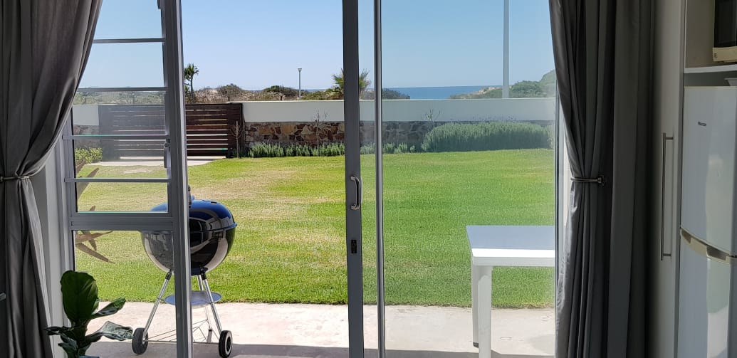 A view from the apartment's sliding glass doors reveals a grassy outdoor space and the ocean in the distance. A Weber grill is situated beside a white table, perfect for outdoor dining. The scene is framed by light curtains, enhancing the inviting atmosphere.