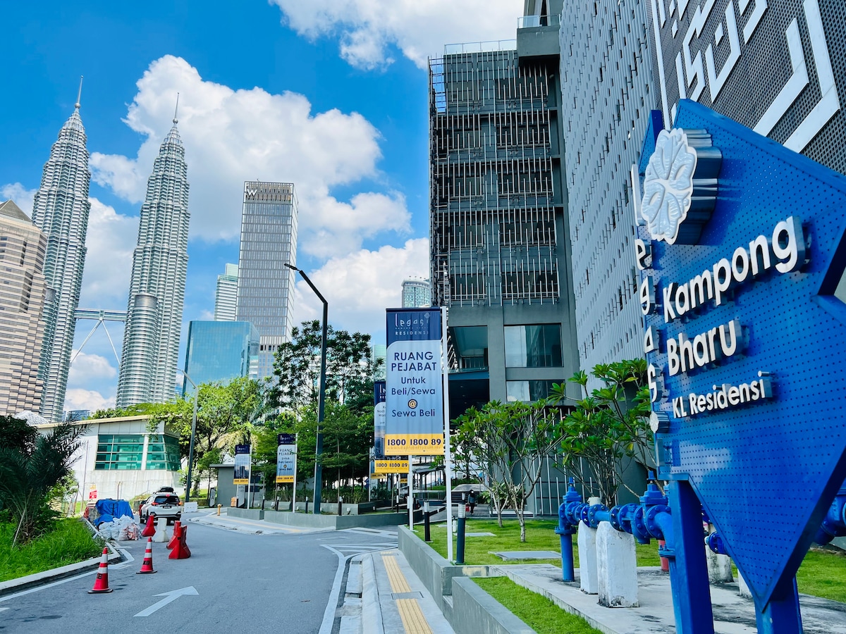 The image showcases a vibrant street scene in Kuala Lumpur's Kampong Bharu. Tall modern buildings, including the iconic Petronas Twin Towers, rise in the background. A clear blue sky and scattered clouds add to the lively atmosphere. Signage and landscaped greenery are visible along the street.