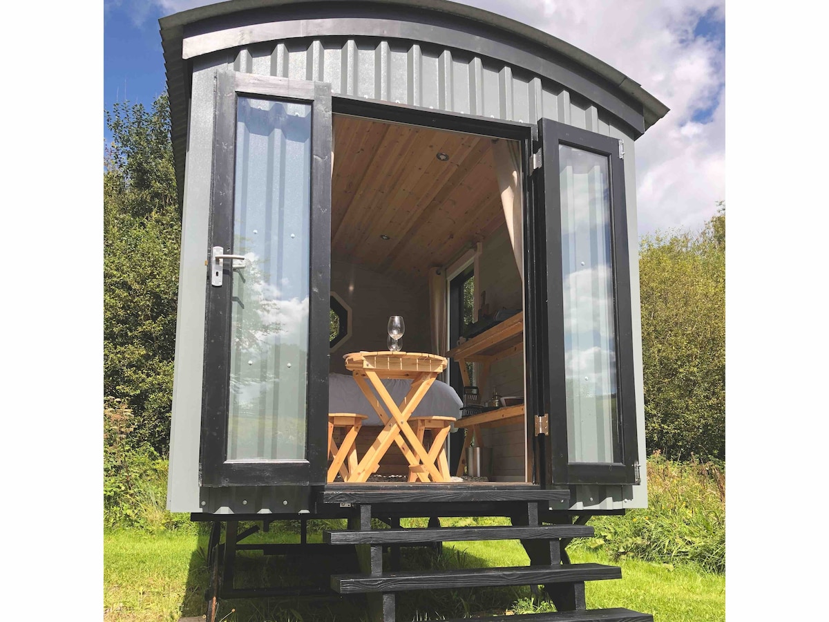 A charming hut features large double doors that open to a welcoming interior, revealing a cozy sleeping area with a bed and a wooden table set for two. The design includes a sleek metal exterior and steps leading up to the entrance.