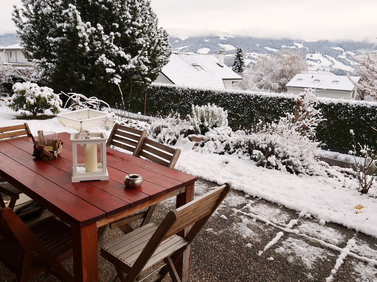 An outdoor patio is presented with a wooden dining table surrounded by several chairs. A lantern and a small bowl decorate the table. A snow-covered landscape is visible in the background, featuring evergreen trees and distant rooftops under a cloudy sky.