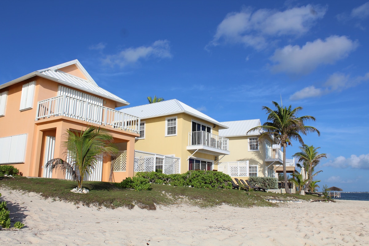 Three beachside villas are shown against a bright blue sky, featuring colorful exteriors in peach and yellow. Lush greenery and palm trees line the sandy shore, inviting relaxation. The structures showcase balconies with white railings, offering access to enjoyable ocean views.