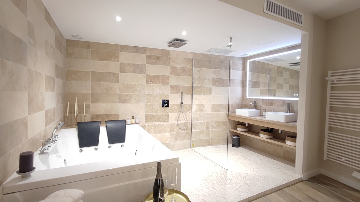 The spacious bathroom features a double whirlpool tub, complemented by two vessel sinks situated on a wooden vanity. A walk-in shower is encased in glass, while soft lighting illuminates the neutral tile walls and floor, creating a calming environment.