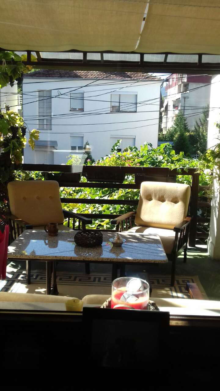 A terrace is shown with two cushioned chairs and a glass table, inviting relaxation. Lush greenery and a view of neighboring buildings are visible in the background, providing a serene outdoor space. Natural light enhances the setting, creating a tranquil atmosphere.
