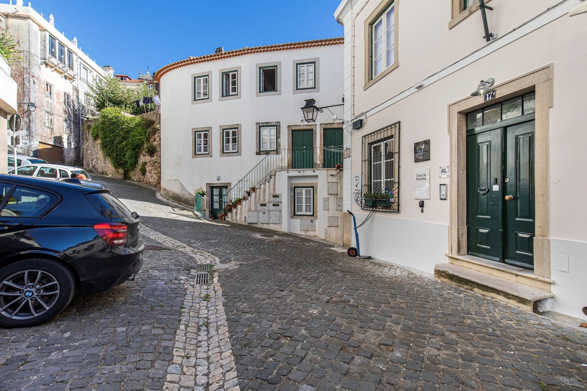 A charming street scene in Sintra features curved white buildings with traditional architecture. Stone-paved roads lead to a set of stairs flanked by greenery and a green door. A parked black car adds to the tranquil urban atmosphere.