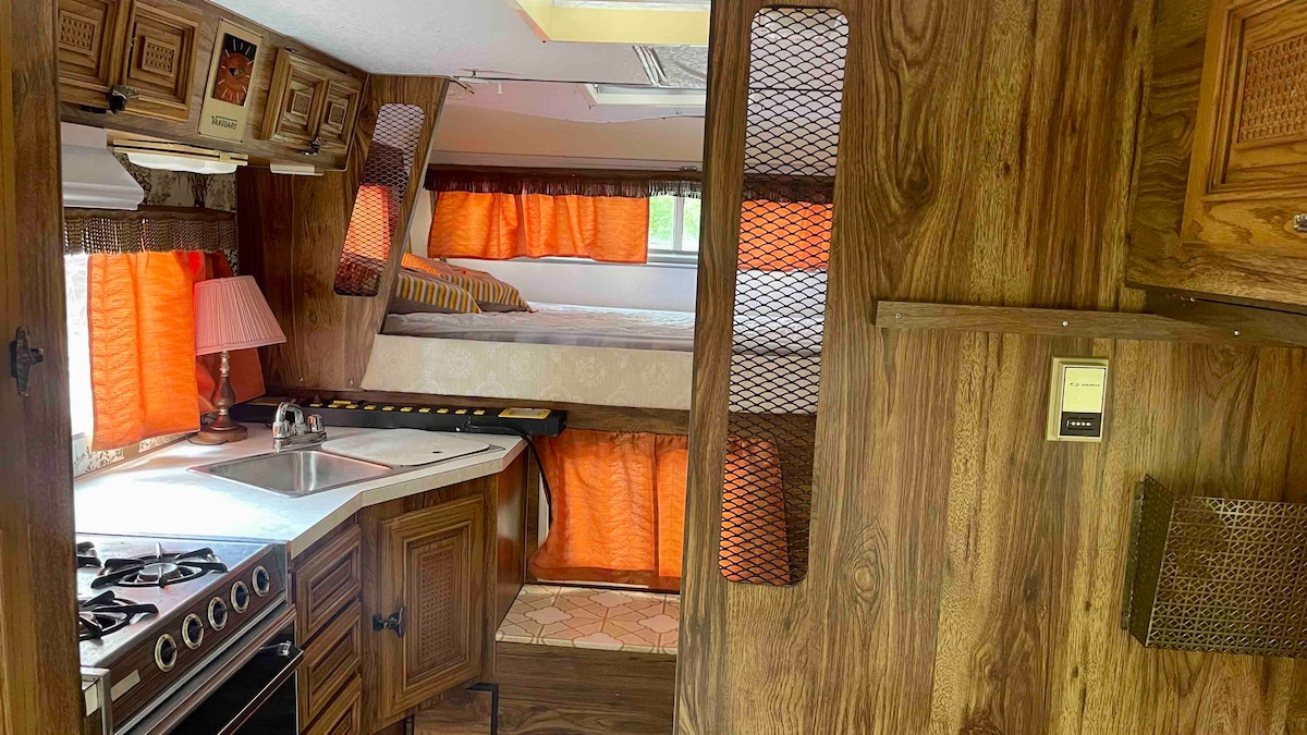 The interior of the camper features wooden cabinetry and warm tones, with a small kitchen area that includes a sink and cooking space. Orange curtains frame the windows, allowing natural light to brighten the space. A bunk bed area is visible in the background, providing additional sleeping arrangements.