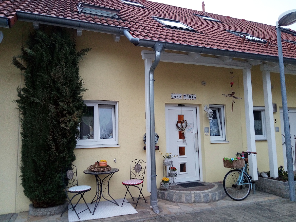 The entrance of Casa Maria features a light yellow facade with decorative elements. A small table and two chairs are positioned outside, complemented by planters and a bicycle. Two windows provide natural light, while the welcoming front door is adorned with a unique design.