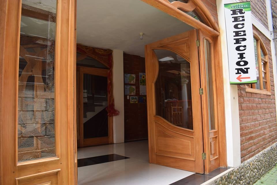 The entrance features large, wooden doors that open to a bright reception area. A polished floor displays contrasting black and white tiles. Artwork is visible on the walls, and an inviting atmosphere is created by the natural light streaming in.