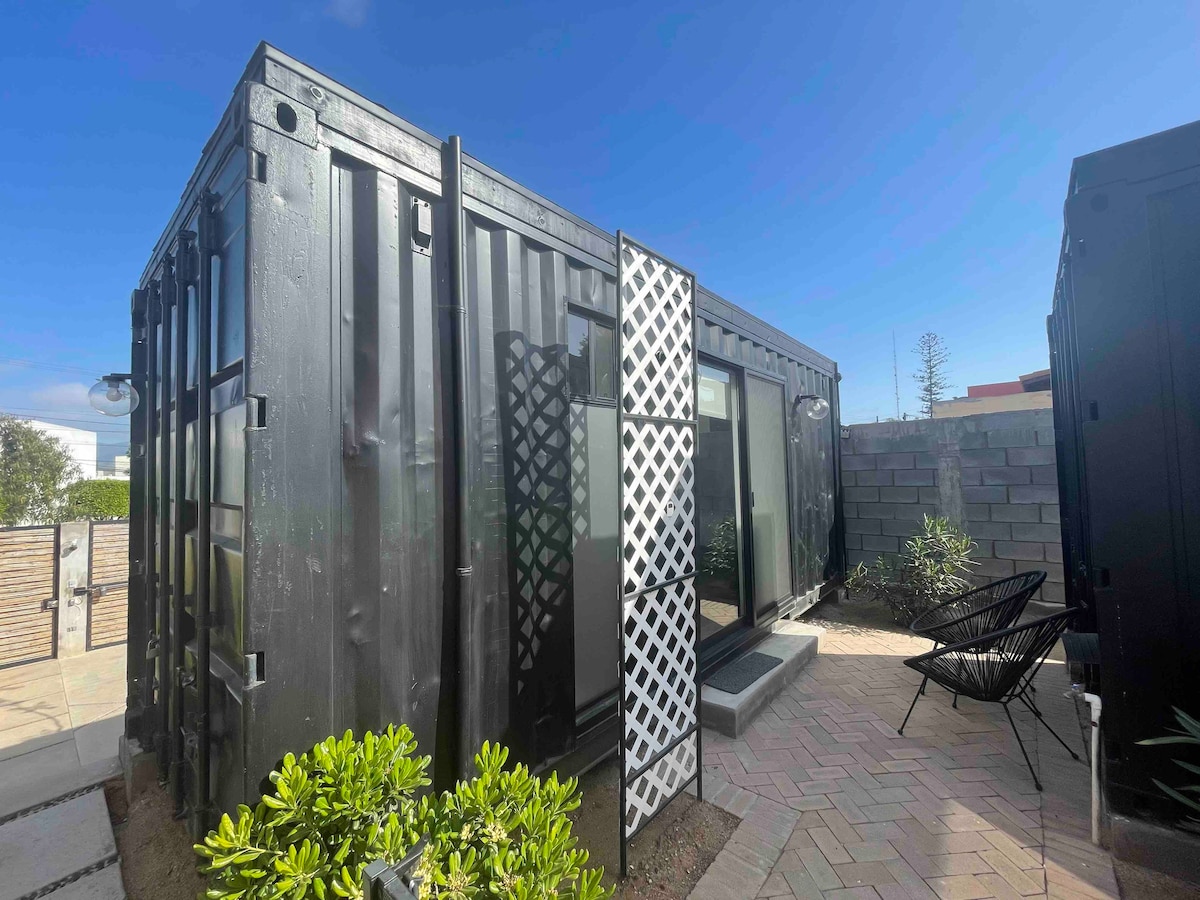 The exterior of the Container Loft is revealed, showcasing a modern design with black walls and large glass doors. A white lattice panel adds visual interest, while a paved area and surrounding greenery create a serene outdoor space, inviting relaxation and enjoyment.