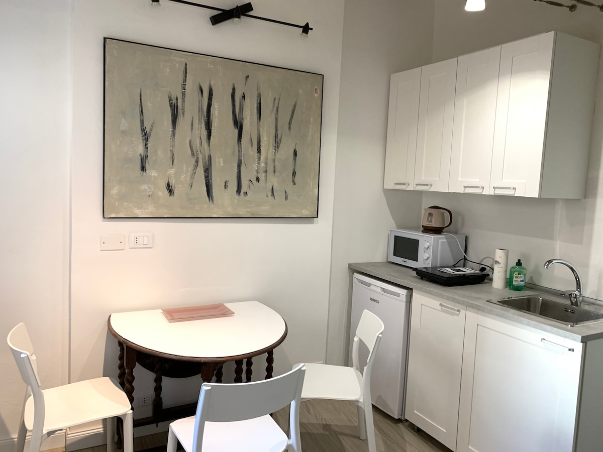 A small kitchen area features a round table surrounded by four chairs. A minimalist artwork hangs above the table. To the side, a microwave and a compact refrigerator are present, alongside a sink and countertop, all designed for efficient use of space.