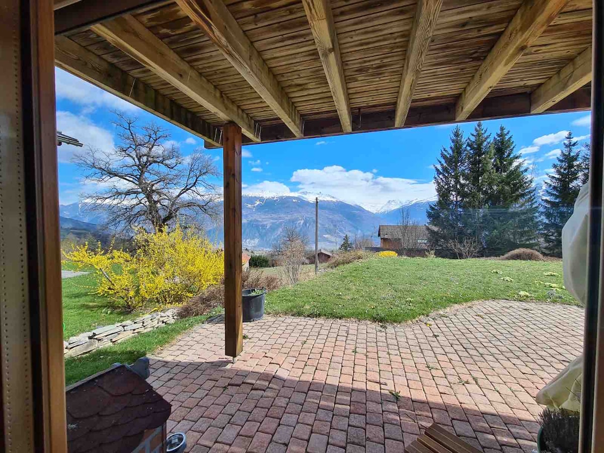 The outdoor patio area features braided stone flooring, leading to a lush green lawn with flowering bushes. A panoramic view of the surrounding mountains is framed by wooden beams overhead. Trees are visible in the background, enhancing the tranquil setting.