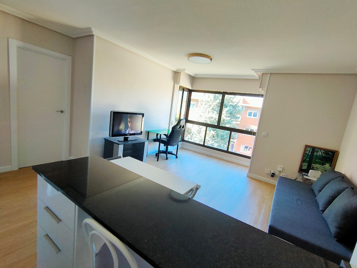 A modern living space is captured, featuring a sleek dark sofa, a television on a low stand, and a desk chair facing a large window. Natural light illuminates the room, highlighting the wooden flooring and spacious layout.