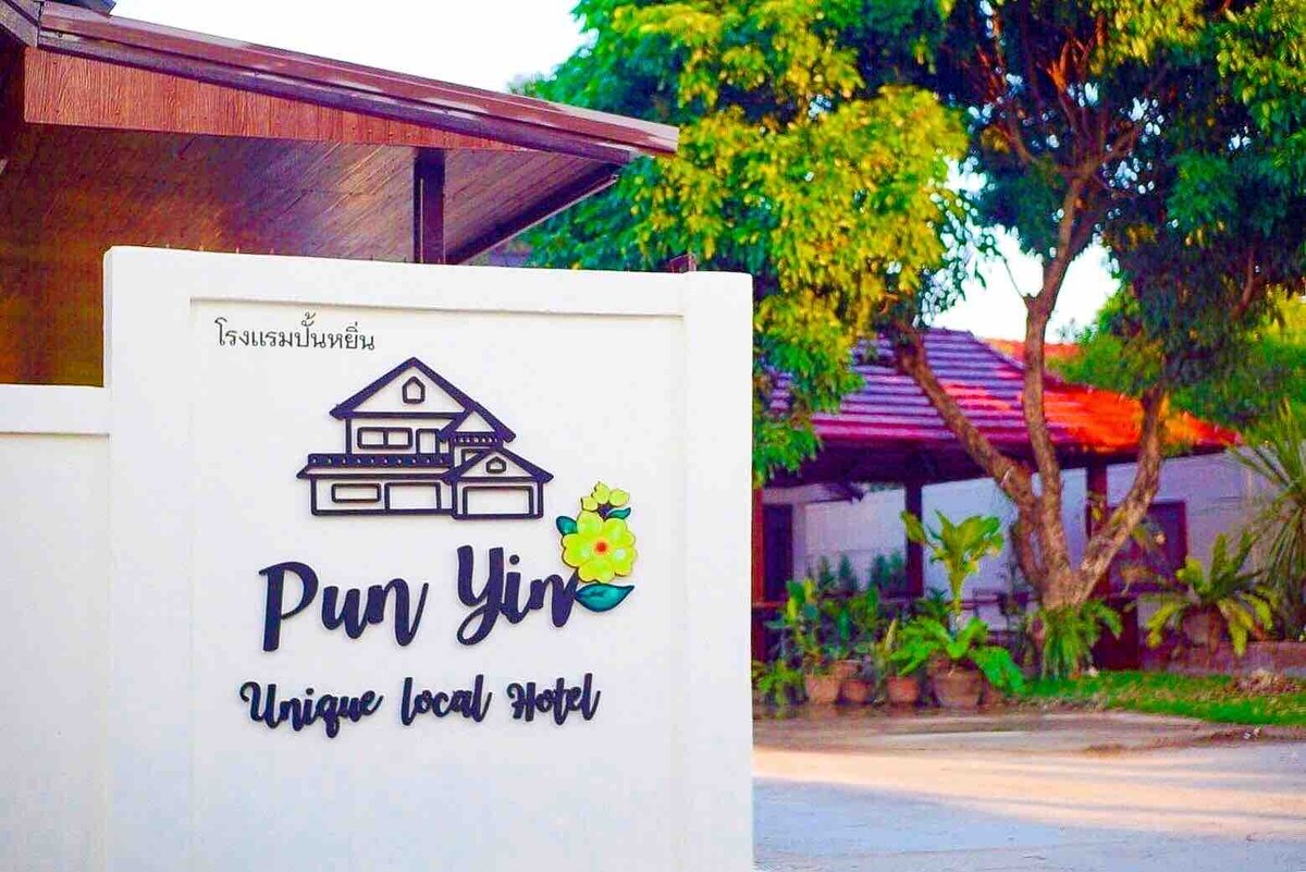 An entrance to a local hotel is shown, featuring a stylish sign with the name 'Pun Yim' and the phrase 'Unique Local Hotel.' Lush greenery surrounds the entrance, and a traditional architecture style is visible in the background.