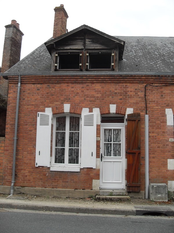 The exterior of the building features traditional red brick walls and white decorative shutters. A panel door with a glass insert is present beneath a small, unadorned window at the top. The roof displays a simple design with exposed timber framing.