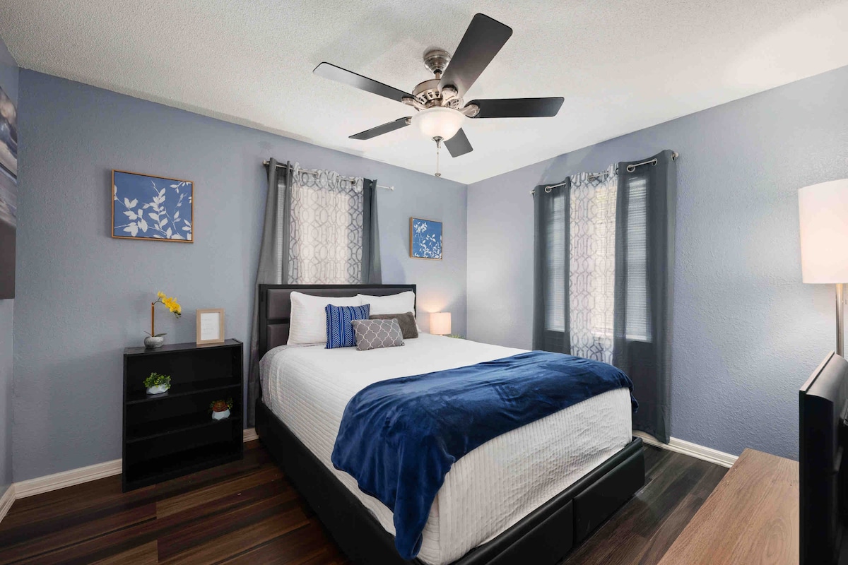 A bedroom is presented with a spacious bed covered in white linens and a blue throw blanket. Two windows adorned with light-filtering curtains allow soft light to enter. A ceiling fan provides air circulation, while a small bookshelf and lamp complete the inviting space.