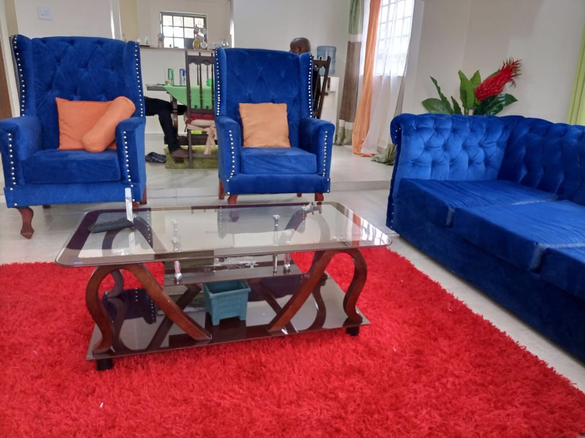 A bright and spacious living area features two blue upholstered armchairs with decorative nailhead trim and an inviting blue sofa. A glass coffee table rests on a vibrant red area rug, while a splash of greenery is provided by potted plants in the background.