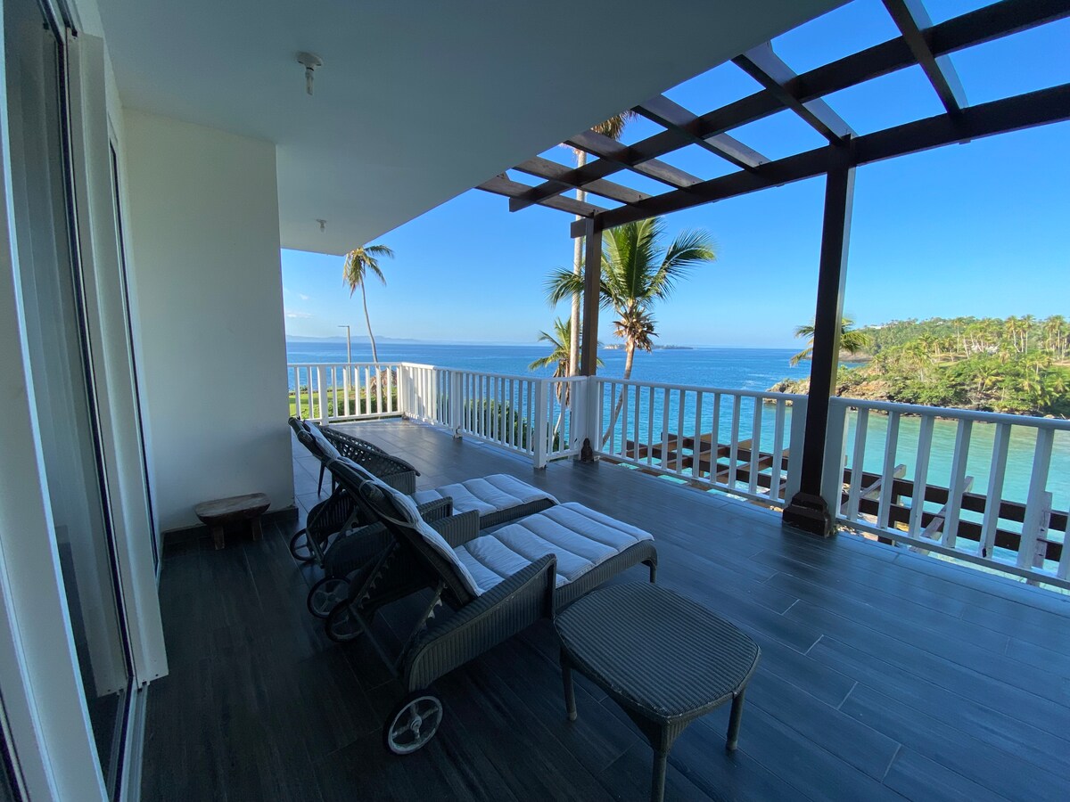 A spacious balcony features several lounge chairs arranged for relaxation, overlooking a stunning coastline. Palm trees gently sway in the breeze, framed by the expansive blue sky. The area is designed for enjoying the peaceful sea views.