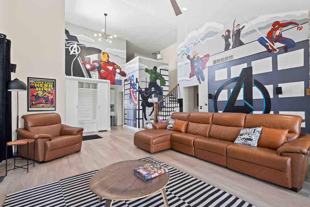 A spacious living room is adorned with Marvel-themed murals depicting various superheroes. A large brown sectional sofa is positioned in front of a round coffee table, while a striped area rug enhances the room's design. Natural light fills the space from tall windows and ceilings.