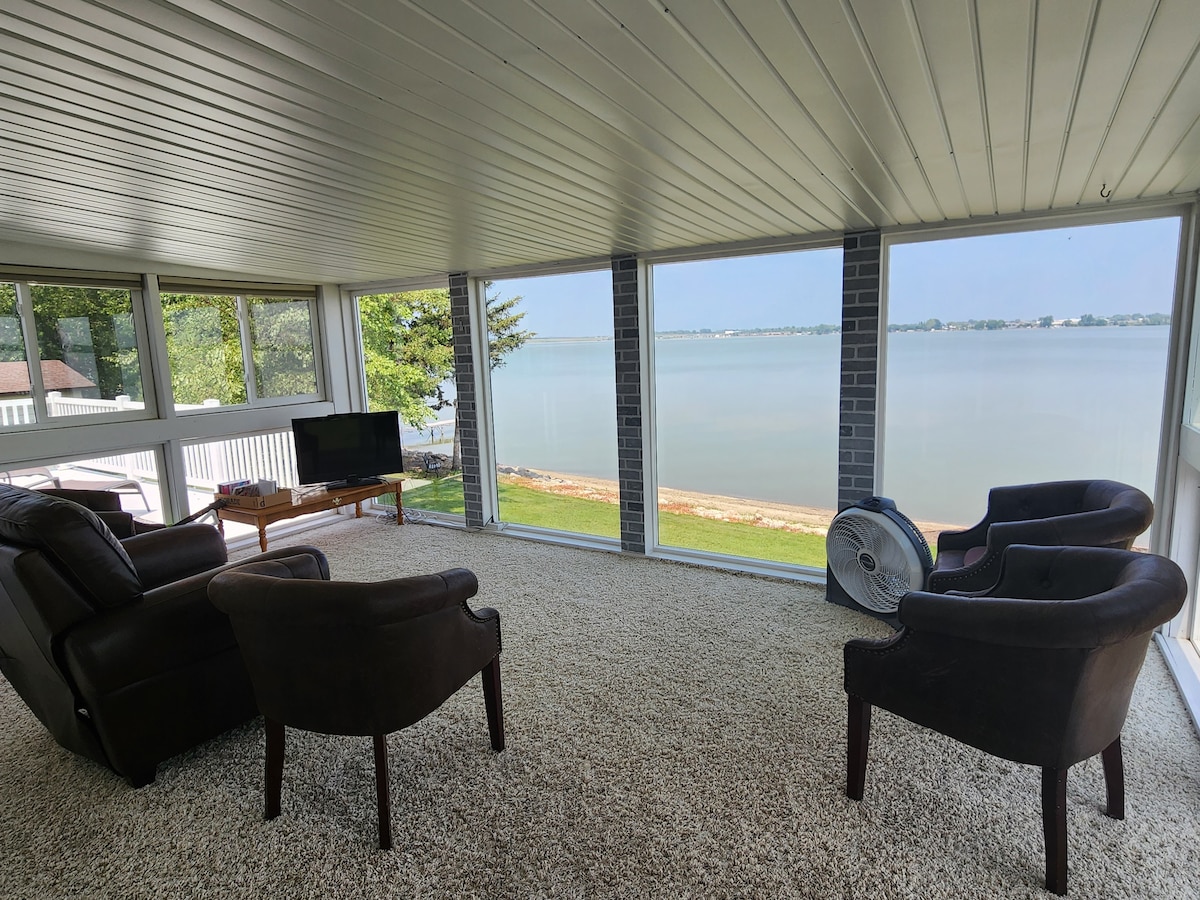 A serene three-season room is filled with natural light from floor-to-ceiling windows, showcasing views of the lake. Two plush chairs and a cozy couch provide seating, while a television sits on a wooden stand. A fan rests on the floor, enhancing comfort.