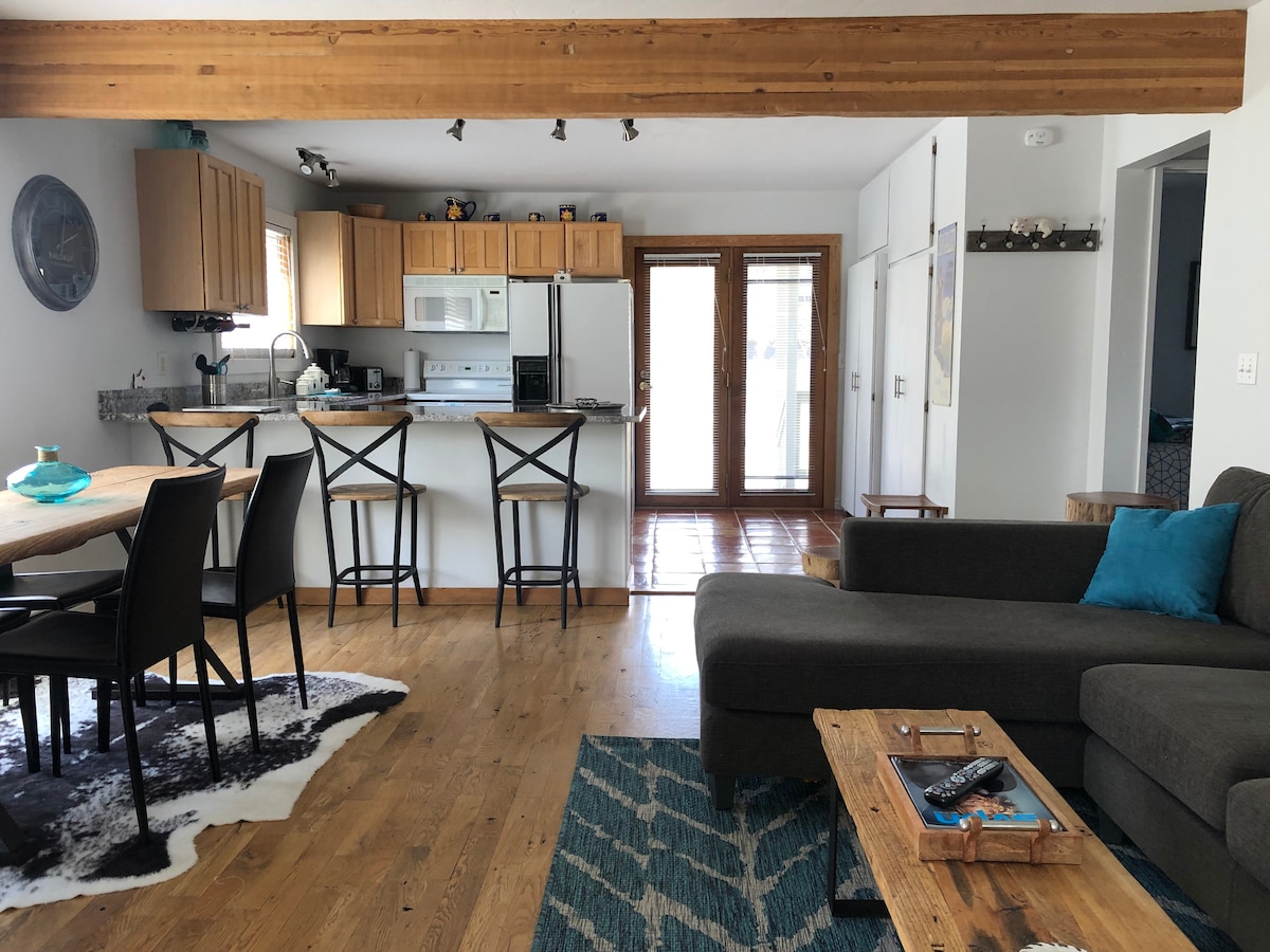 An open living area features a comfortable sectional sofa facing a wooden coffee table. A dining table with chairs is set nearby, and a kitchen bar with stools adds to the space. Natural light floods in from the glass doors leading to the porch.