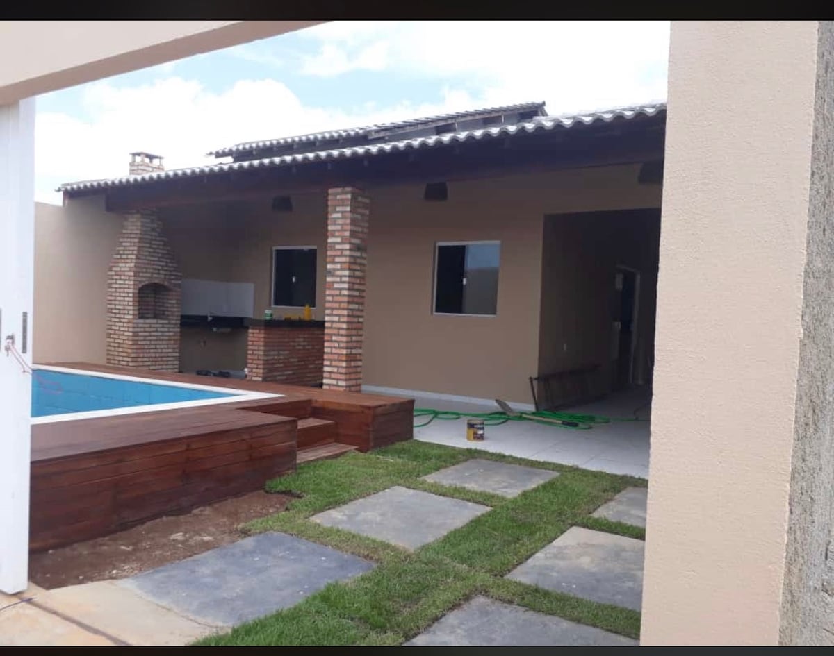 An outdoor area features a pool with wooden decking, accompanied by a grill station made of brick. The structure showcases an inviting facade with large windows and a spacious layout, while the surrounding grass and stone pathway add a touch of greenery.