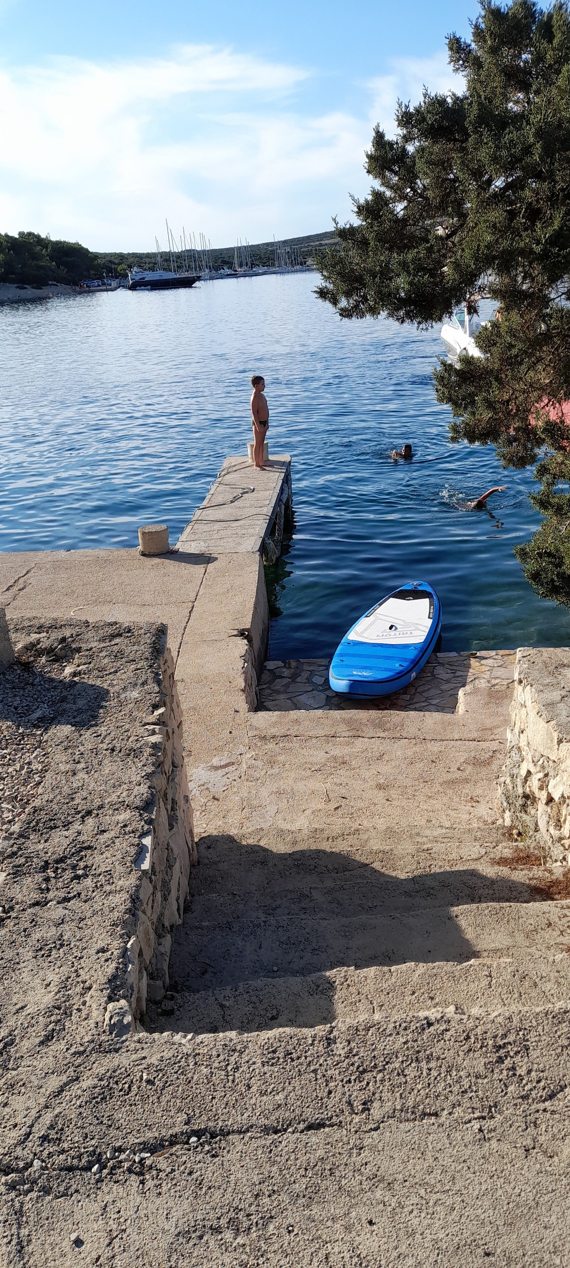 A stone jetty extends into calm waters, with a paddleboard resting nearby. Steps lead down to the water's edge, where two individuals can be seen swimming. Lush greenery surrounds the scene, with a boater visible in the distance along the shore.