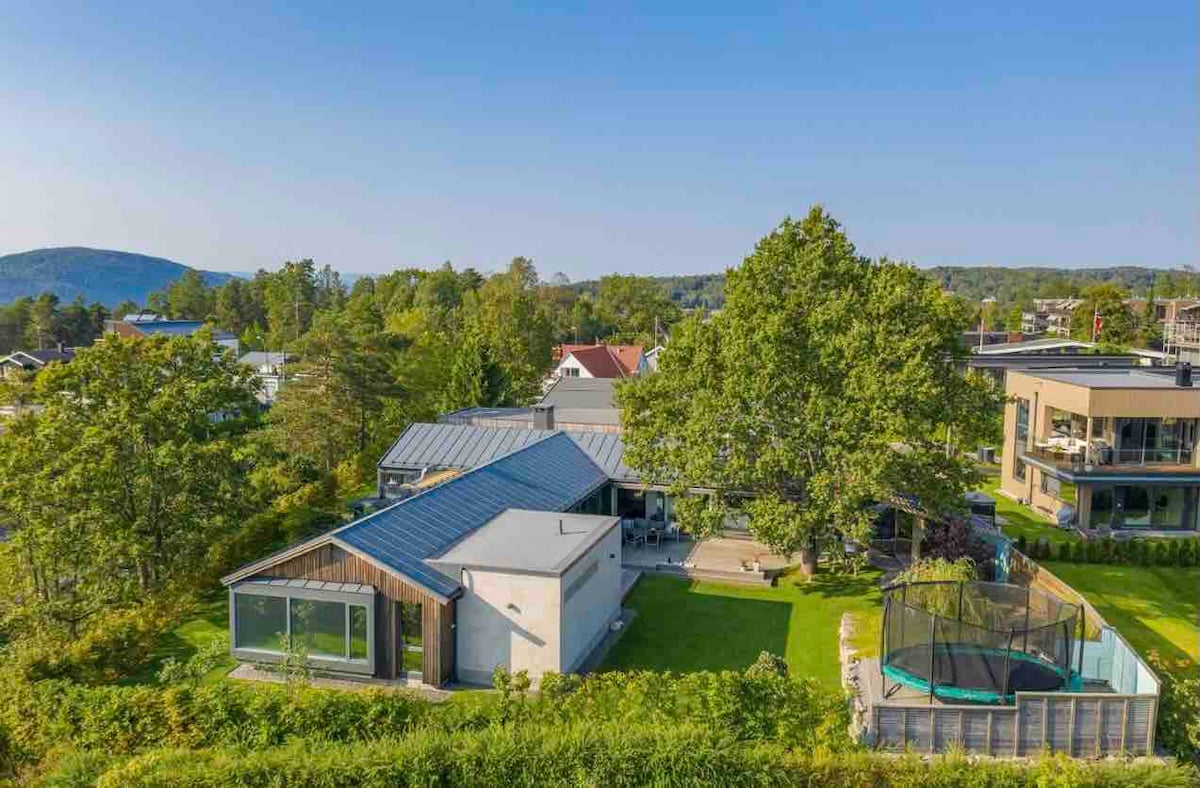 An aerial view of a spacious modern house surrounded by lush greenery, showcasing a large private garden. A trampoline can be seen in the garden area, along with a charming roof that adds character to the structure.