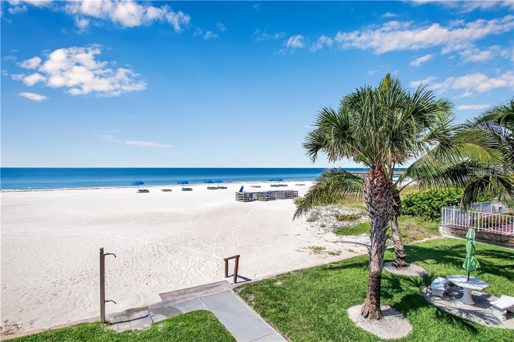 A scenic view of the sandy beach stretches towards the ocean, with lounge chairs arranged on the shore. Lush palm trees frame the scene, while a walkway leads directly to the beach. Clear blue skies and gentle waves create a peaceful coastal atmosphere.