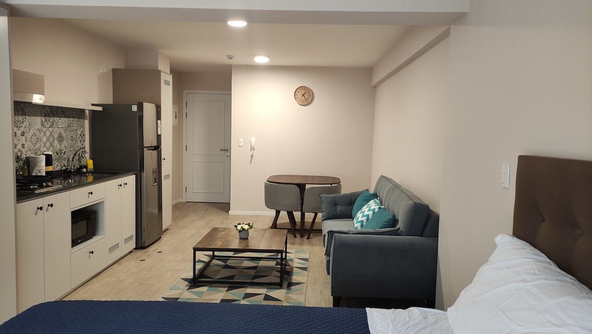 A modern loft interior is displayed, featuring a kitchenette with dark cabinetry and a sleek refrigerator. A dining area with a round table is positioned near a comfortable sofa, which is complemented by a colorful accent pillow. Natural light enters through a nearby doorway.