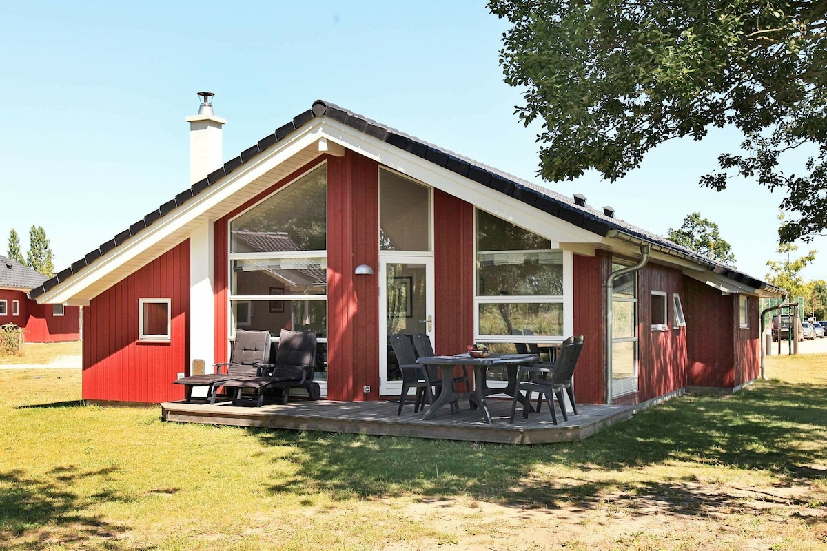 A modern red holiday home is showcased in a landscaped area, featuring large windows and a sloped roof. Outdoor furniture includes a dining table and lounge chairs on a wooden terrace, surrounded by green grass and trees.