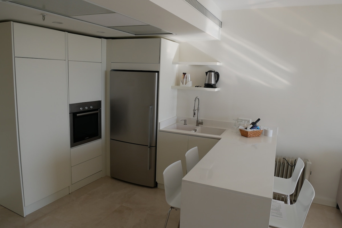 A modern kitchen is equipped with sleek, white cabinetry and stainless-steel appliances. An island with seating for two is complemented by a minimalist design. Above the sink, ample natural light enters through a nearby window, enhancing the space's functionality and openness.