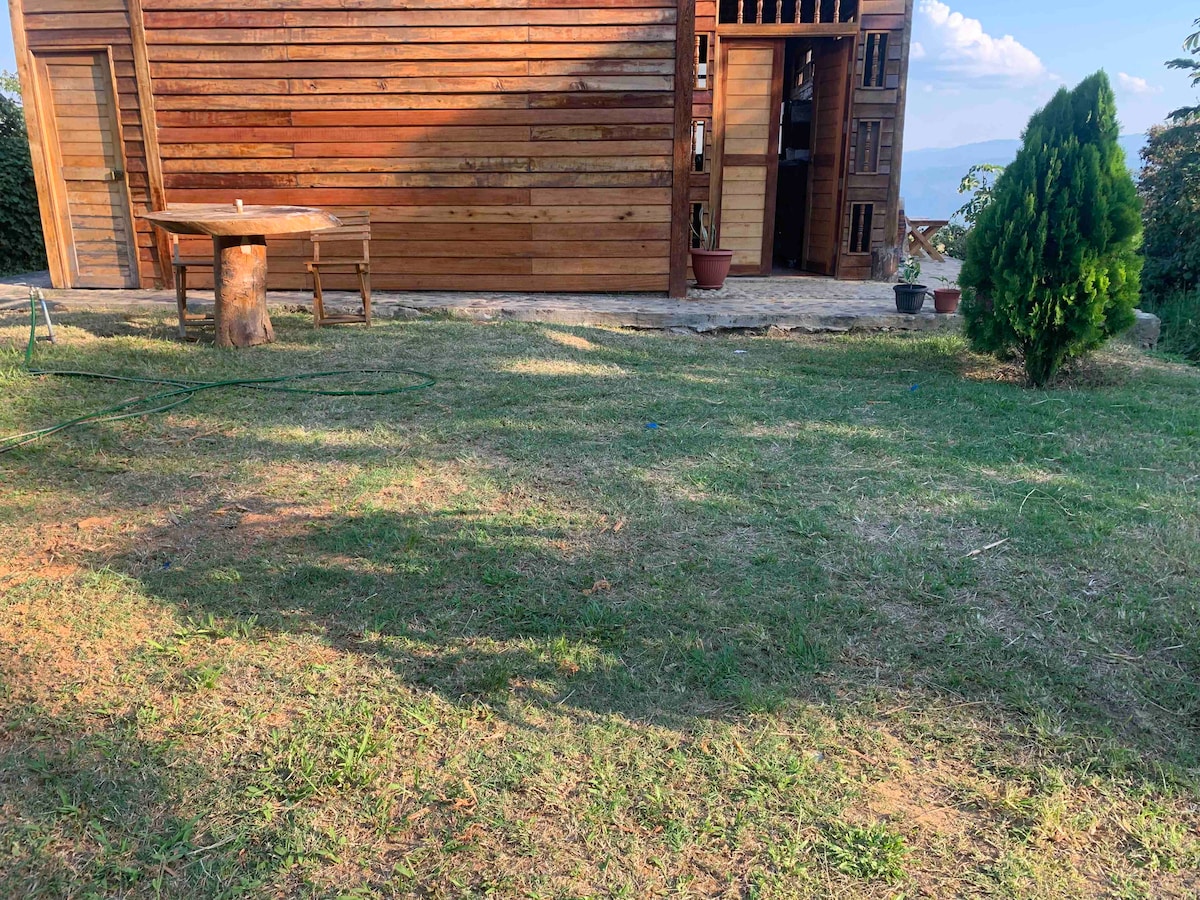 The exterior of the cabin features a wooden façade, complemented by a spacious lawn area. A wooden table and chairs are positioned on the grass, inviting outdoor dining. Potted plants are placed near the entrance, enhancing the connection with nature.