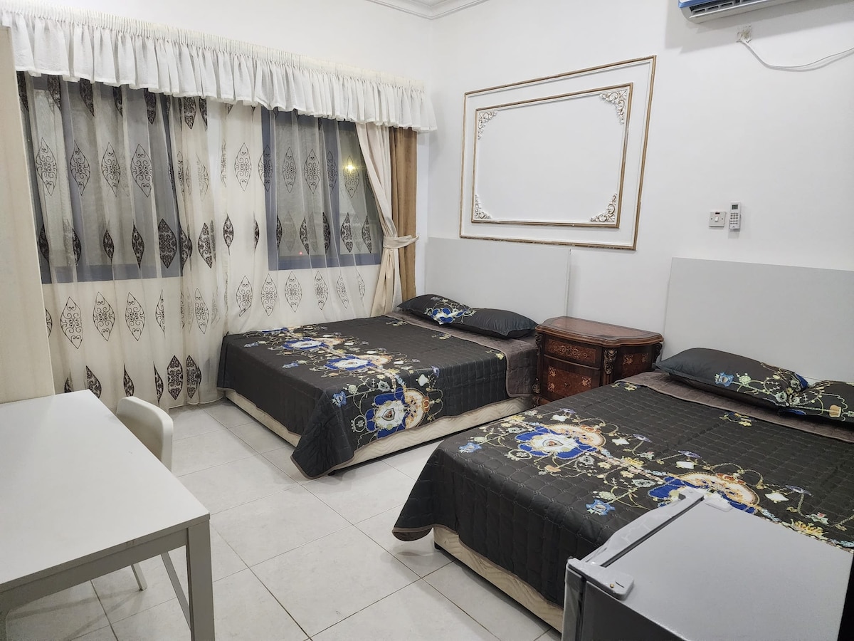 The room features two spacious beds covered with dark, patterned bedding. A wooden cabinet is placed beside the beds, and a small table is positioned near the window, which is dressed with light, patterned curtains. A compact refrigerator is also visible in the corner.