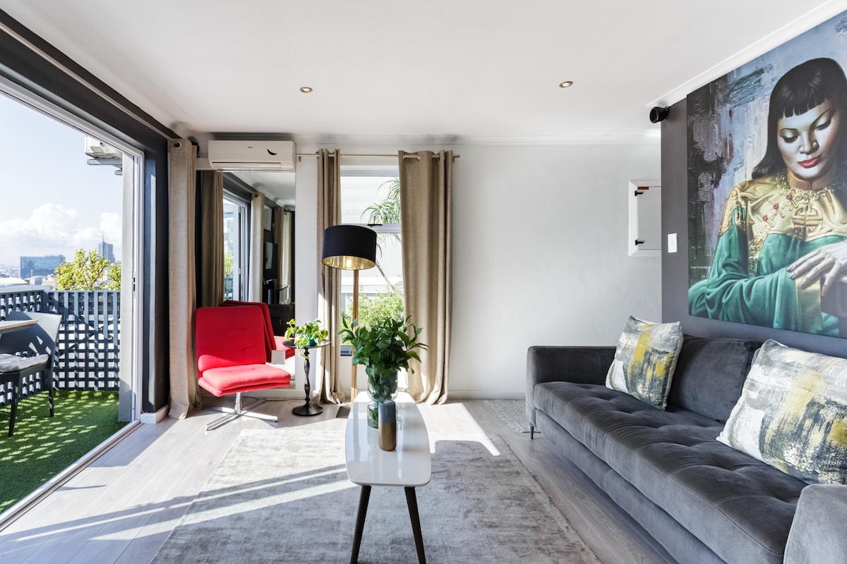 A bright living area is depicted, featuring a gray sofa adorned with decorative pillows. Natural light fills the space, highlighted by large sliding doors that provide access to an outdoor balcony. A stylish lamp stands next to a red accent chair, with greenery visible in the background.