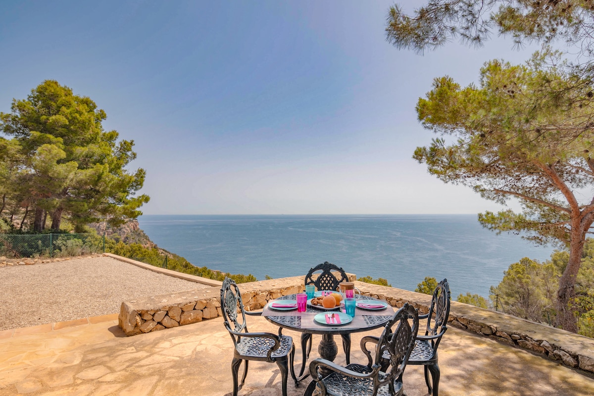 An outdoor dining area features a round table surrounded by black wrought iron chairs, set against an expansive view of the sea. A variety of colorful tableware is arranged atop the table, with trees and a serene landscape visible in the background.