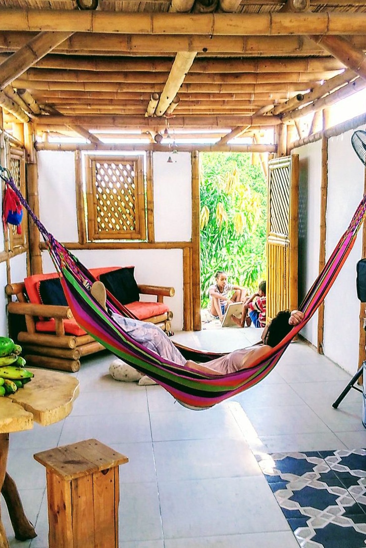A colorful hammock is suspended in a cozy living area with wooden accents. Natural light filters in through open doors, illuminating a casual seating arrangement and a small dining table. Lush greenery is visible outdoors, enhancing the connection with nature.
