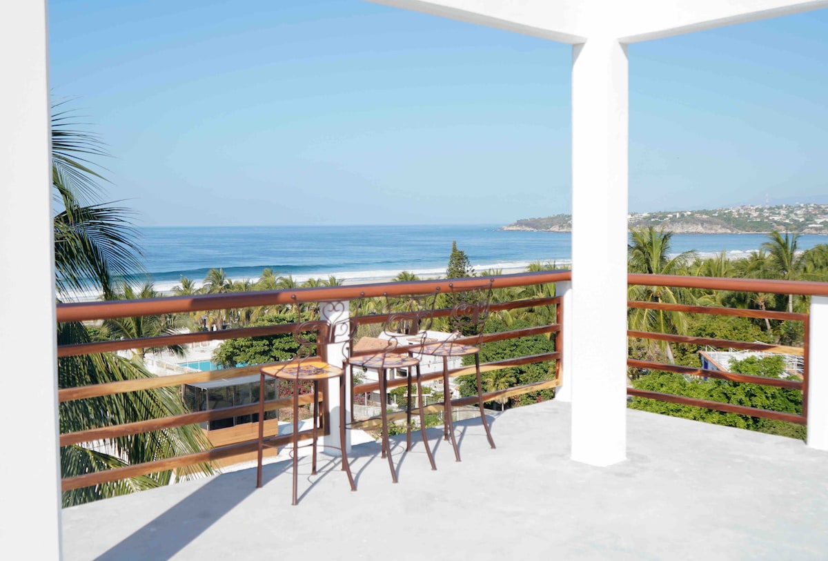 A private balcony offers a stunning view of the ocean, framed by lush palm trees. Two wooden chairs and a small table are positioned to enjoy the scenery, with the coastline visible in the distance under clear blue skies.