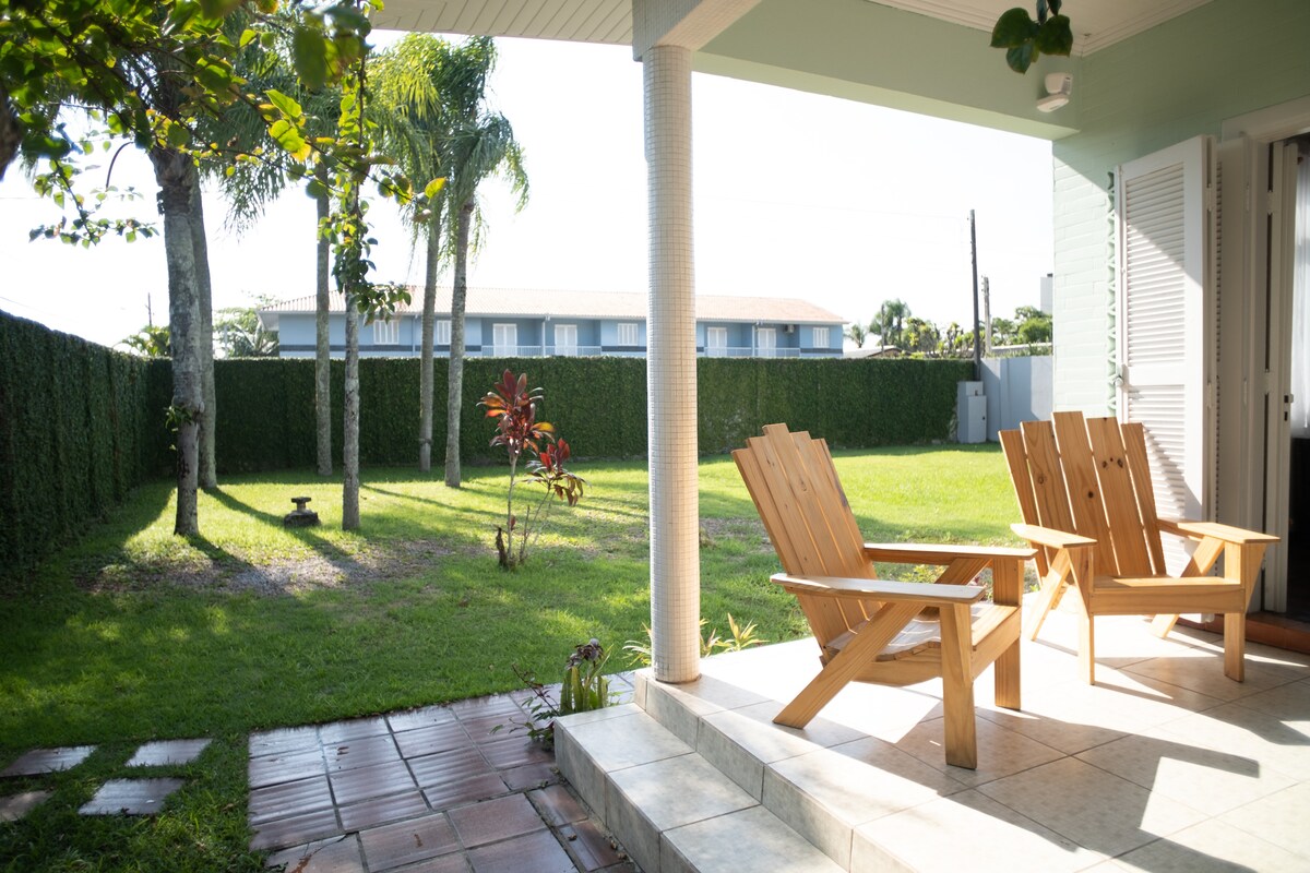 Two wooden chairs are positioned on a tiled porch, offering a view of the expansive green yard. Sunlight brightens the scene, highlighting the lush grass and surrounding trees. A small garden area is visible in the background, providing a tranquil setting.