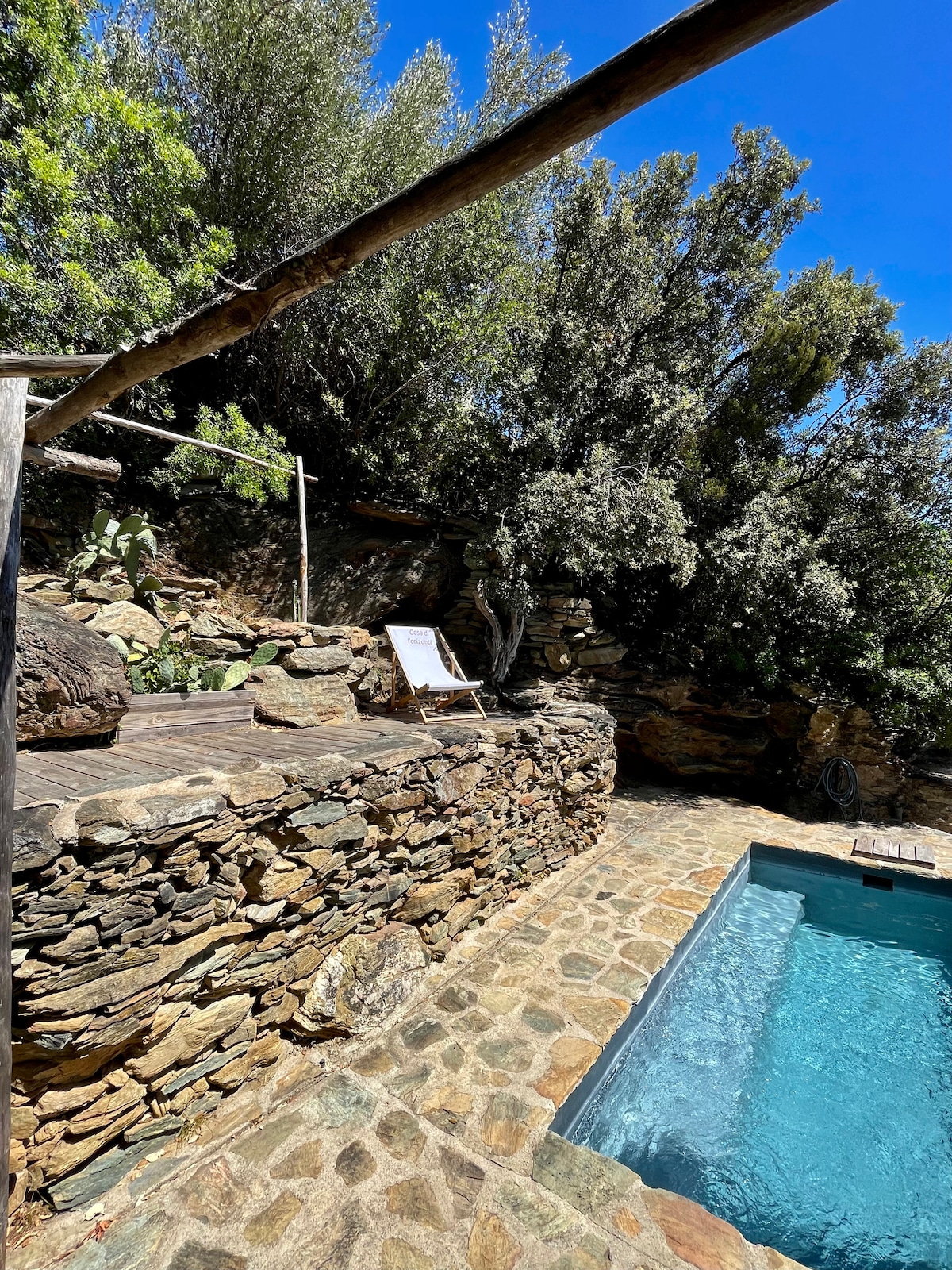 A traditional Corsican pool area is framed by natural stone walls, integrated with the landscape. Sun loungers are positioned beside the water, surrounded by lush greenery. The blue water reflects the clear skies, creating a serene outdoor space for relaxation.