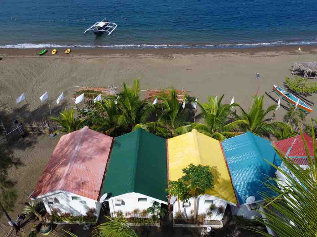 Aerial view of several charming tiny homes, featuring roofs in vibrant colors including red, green, yellow, and blue. The beachfront setting is complemented by sandy shores and palm trees, creating a serene atmosphere. A boat can be seen on the calm water, enhancing the coastal ambiance.