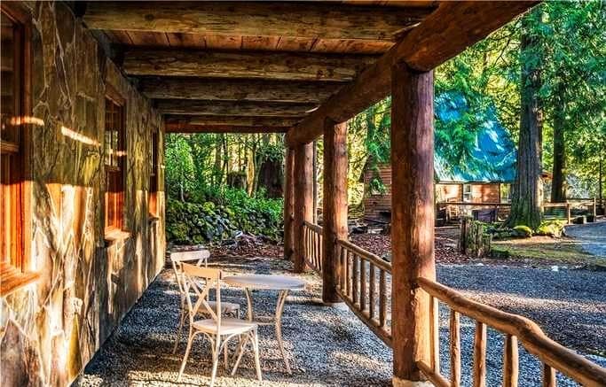 A log cabin porch features a wooden ceiling and rustic posts, with soft sunlight filtering through the trees. A small round table with two chairs is placed on the gravel surface, surrounded by natural scenery, inviting peaceful moments outdoors.