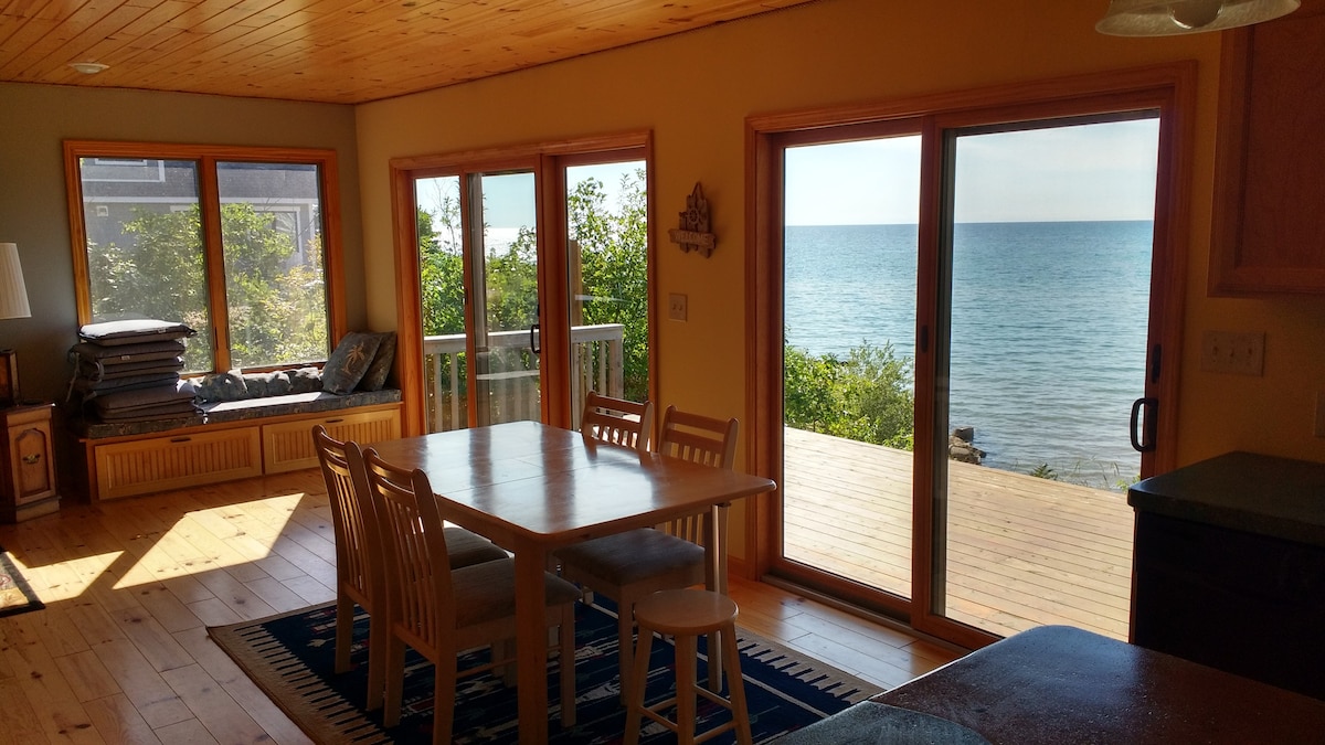 A spacious dining area features a wooden table surrounded by six chairs, with a vibrant area rug beneath. Large windows offer views of Lake Michigan, bringing natural light into the room. A cozy seating nook with pillows is visible beside the window.