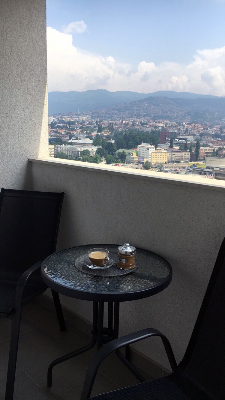A small balcony features a black glass table with a cup of coffee and a sugar container. Two matching chairs provide seating. The expansive view showcases the cityscape and surrounding mountains under a partly cloudy sky.