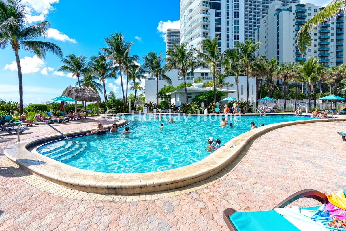 The pool area features a large, inviting swimming pool surrounded by lush palm trees. Lounge chairs line the pool, with umbrellas providing shade. Guests can be seen enjoying the water and relaxing by the poolside, creating a lively atmosphere.