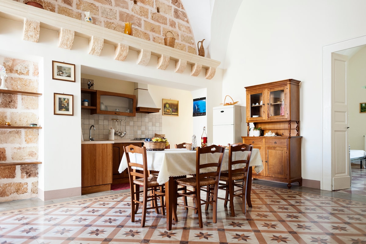 A spacious kitchen area features a wooden dining table surrounded by chairs, complemented by traditional stone flooring. A well-equipped kitchen is visible, with wooden cabinetry and modern appliances. Natural light illuminates the space, enhancing the warm tones of the decor and the inviting atmosphere.